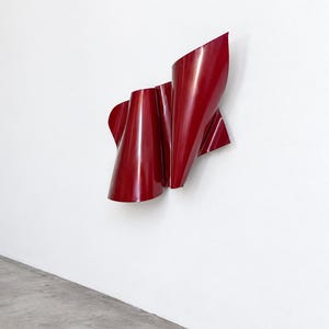 May include: A deep red wall sculpture featuring a series of smooth, curved, and folded metallic panels. The object has a glossy, reflective finish and an abstract, undulating form that creates sharp edges and deep shadows across its surface.