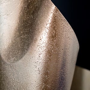 May include: A curved sheet of metallic bronze material with a distinctively rough, granular, and textured surface. The light reflects off the uneven, sandy-like finish, creating highlights and shadows that emphasize the material's rigid, curved form.