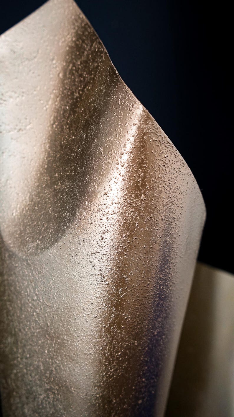 May include: A curved sheet of metallic bronze material with a distinctively rough, granular, and textured surface. The light reflects off the uneven, sandy-like finish, creating highlights and shadows that emphasize the material's rigid, curved form.
