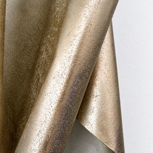 May include: A metallic gold sheet with a heavily textured, granular surface that curves and folds over itself. The material displays a shimmering, uneven finish with darker bronze undertones visible in the recessed areas of the textured pattern.