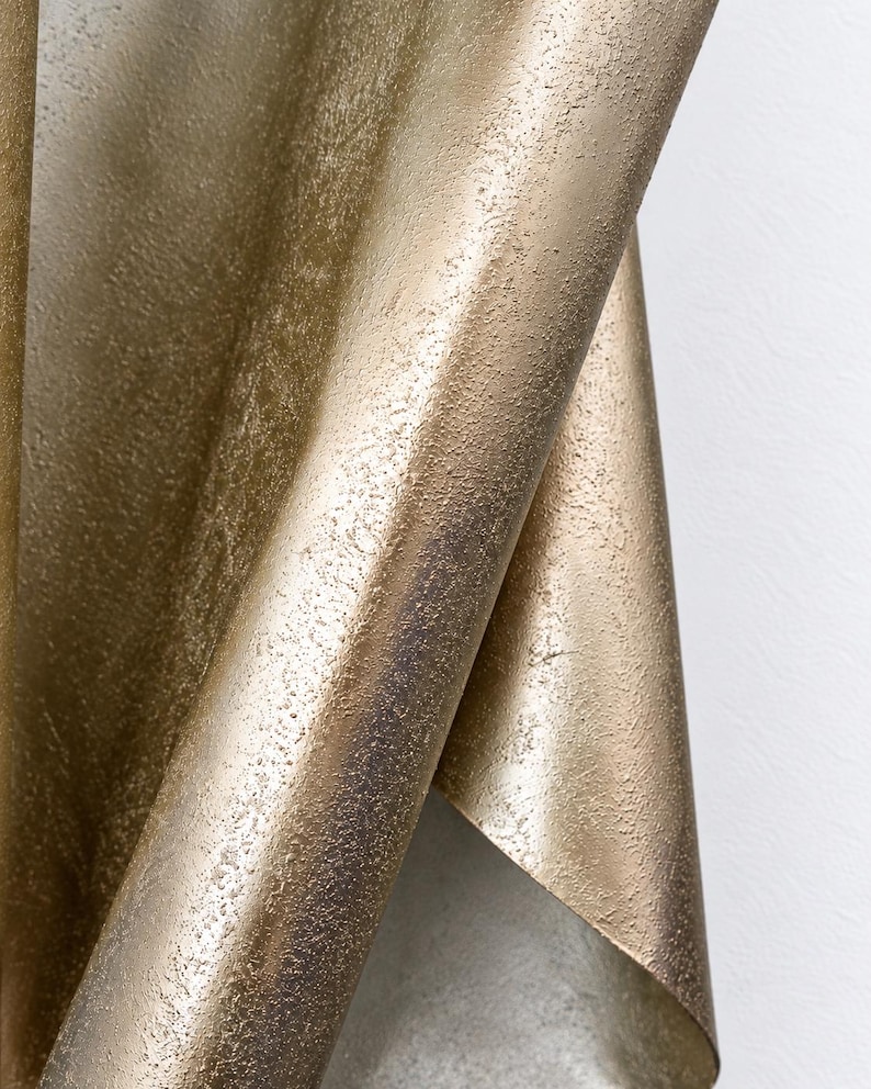 May include: A metallic gold sheet with a heavily textured, granular surface that curves and folds over itself. The material displays a shimmering, uneven finish with darker bronze undertones visible in the recessed areas of the textured pattern.
