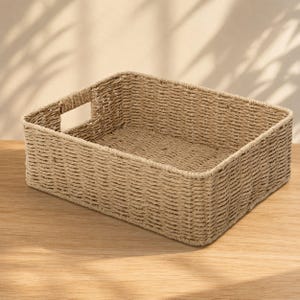 May include: A rectangular storage basket crafted from woven light brown natural fibers. The basket features a structured open-top design with integrated cutout handles on two opposing sides, showcasing a consistent braided texture throughout.