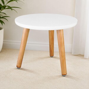 May include: A round side table featuring a smooth white circular tabletop supported by three tapered light brown wooden legs. Each leg is finished with a small metallic circular foot pad at the base, providing stability for the furniture piece.