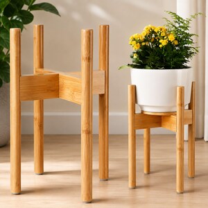 May include: Two light brown bamboo plant stands with a cross-shaped support structure and four cylindrical legs. One stand is empty, while the other holds a white ceramic pot containing a green plant with small yellow flowers.