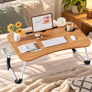 Puede incluir: Una mesa de madera para portátil con patas plegables blancas y pies negros antideslizantes. La mesa incluye una ranura para tableta, un portavasos y un cajón. Sobre la superficie hay una tableta, un teclado blanco, un ratón, un cuaderno y una taza.