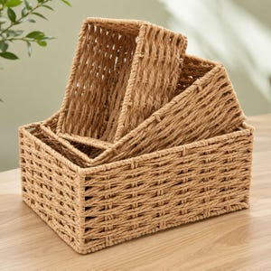 May include: A set of three rectangular storage baskets crafted from light brown woven material. The baskets are nested together, featuring an open-top design with a consistent grid-like weave pattern throughout the sides and base of each unit.