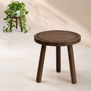 May include: A small, round, dark brown wooden stool with three cylindrical legs. The top surface features a circular recessed groove near the edge. The stool is constructed from solid wood with a smooth, polished finish and a sturdy, stable design.