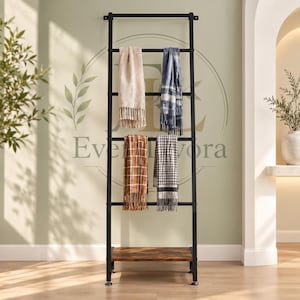 May include: A tall black metal ladder rack with five horizontal rungs and a wooden base shelf. Four folded blankets with different patterns and colours, including beige, blue, brown, and grey, are draped over the rungs of the rack.