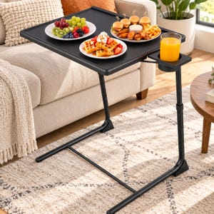 May include: A black adjustable TV tray table with a rectangular surface and a side cup holder. The table holds three white plates containing waffles with strawberries and whipped cream, assorted fresh fruit, and sliders with french fries.