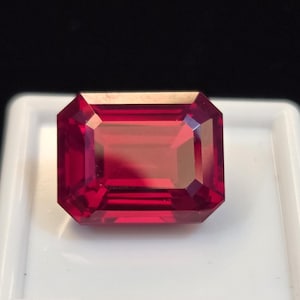 May include: A vibrant, deep red gemstone with a rectangular emerald cut, featuring sharp, clean facets that reflect light across its polished surface. The stone is displayed centered on a square white base, showcasing its rich, saturated color.