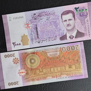Puede incluir: Dos billetes de 2000 libras sirias en color púrpura y blanco. El billete superior presenta un retrato de Bashar al-Assad y el edificio del Banco Central de Siria. El billete inferior muestra el edificio del Parlamento sirio y el texto 2000.