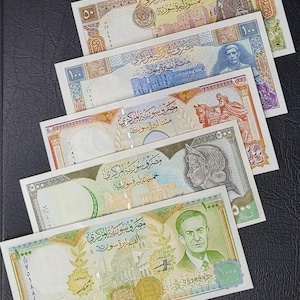May include: A collection of five Syrian banknotes in denominations of 50, 100, 200, 500, and 1000 pounds. Each rectangular paper bill features intricate geometric patterns, Arabic text, and portraits of historical figures or architectural landmarks.