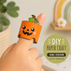 Puede incluir: Un anillo de papel con una calabaza de color naranja brillante con una cara de jack-o'-lantern negra, hojas verdes y un tallo marrón. El texto en la imagen dice DIY PAPER CRAFT RING VERSION DIGITAL DOWNLOAD. El anillo se lleva en un dedo.