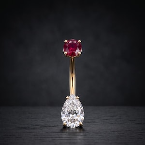 May include: A gold navel piercing ring featuring a round-cut red gemstone at the top and a larger pear-shaped clear gemstone at the bottom, both held in place by four-prong settings on a curved metal barbell.