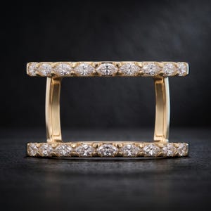 May include: A yellow gold ring guard featuring two parallel bands connected by vertical bars. Each band is set with a row of marquise-cut white diamonds. The design creates an open rectangular space between the two horizontal diamond-encrusted bands.