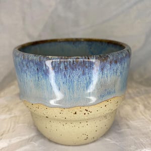 Può includere: Una tazza in ceramica dal design bicolore, caratterizzata da una base color crema maculata e da uno smalto blu lucido e colato sulla parte superiore. Il bordo è marrone scuro e il corpo presenta un rilievo orizzontale che separa le texture.