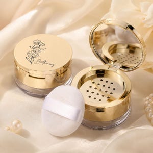 May include: Two round cosmetic powder containers with shiny gold lids and clear bases. One lid features a black floral line drawing and the name Bethany. The other container is open, revealing an internal sifter and a mirror, with a white puff.