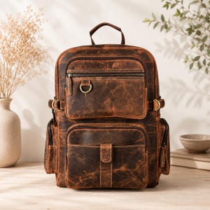 Peut inclure: Un sac à dos en cuir vieilli marron foncé doté d'une poignée supérieure, de deux poches avant zippées, de poches latérales et de ferrures en laiton. La poche avant supérieure inclut un anneau en D et la poche inférieure a un rabat à pression.
