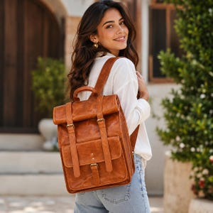May include: A tan leather roll-top backpack featuring two vertical straps with metal buckles, a top handle, and a front pocket with a central buckle closure. The bag has a smooth, worn-in texture and is worn on the back with adjustable shoulder straps.