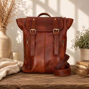 Peut inclure: Un sac à dos en cuir couleur cognac doté d'un rabat supérieur repliable, sécurisé par deux sangles verticales avec des boucles en métal doré. Le sac comprend deux poches latérales zippées, une poignée supérieure et une bandoulière réglable.