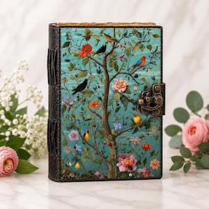 Könnte beinhalten: Ein rechteckiges Notizbuch mit schwarzem Lederrücken und einem Vorderdeckel, der einen türkisfarbenen Hintergrund mit einem Baum, bunten Vögeln und verschiedenen Blumen zeigt. Das Notizbuch hat einen dekorativen Verschluss aus Messing.
