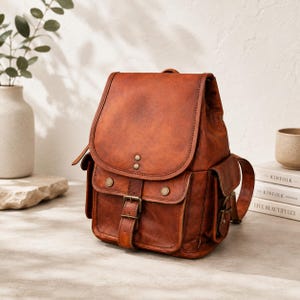 May include: A cognac-coloured leather backpack featuring a fold-over flap with two brass-toned rivets, a front pocket secured by a buckle strap, and two side pockets with brass-toned snap closures. The bag has visible stitching and a smooth finish.