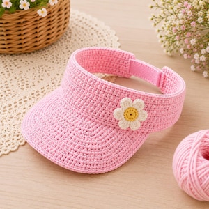 May include: A bright pink crocheted sun visor featuring a single white and yellow daisy appliqué on the front. The visor has a textured, woven appearance with a hook-and-loop closure strap at the back to adjust the fit securely around the head.
