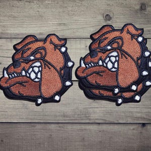 Può includere: Due toppe ricamate raffiguranti la testa di un bulldog con pelo marrone, contorni neri e denti bianchi. Il design include un collare nero decorato con punte bianche. Le toppe sono identiche per colore, forma e cuciture dettagliate.