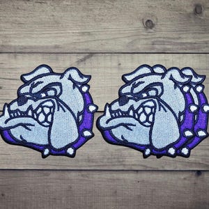 Pode incluir: Dois emblemas bordados com a cabeça de um bulldog com pelo azul claro, contornos azul escuro e uma coleira roxa adornada com espinhos brancos. Os emblemas exibem uma expressão facial estilizada com dentes e orelhas visíveis.