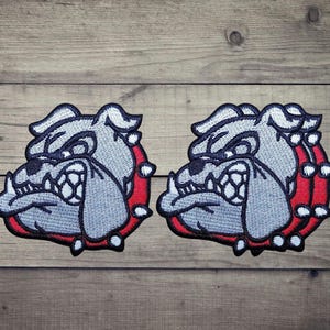 Può includere: Due toppe ricamate identiche raffiguranti una testa di bulldog stilizzata nei toni del grigio, nero e bianco. Il bulldog è raffigurato con un muso prominente, la fronte corrugata e un spesso collare rosso ornato di punte bianche sul collo.