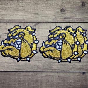 Può includere: Tre toppe ricamate con un design stilizzato di testa di bulldog. Ogni toppa mostra un volto giallo con contorni blu scuro, denti bianchi e accenti bianchi. Il bulldog ha la fronte aggrottata e un'espressione ringhiante con zanne visibili.