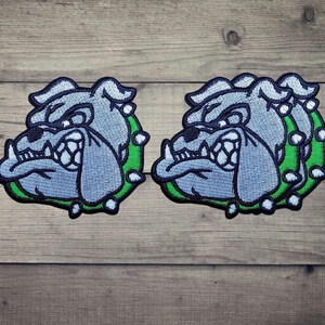 Könnte beinhalten: Zwei gestickte Aufnäher mit einem stilisierten Bulldoggenkopf in Hellblau und Weiß, schwarz umrandet, mit einem leuchtend grünen Halsband mit weißen Stacheln. Das Design ist bei beiden Aufnähern identisch und zeigt einen grimmigen Gesichtsausdruck.