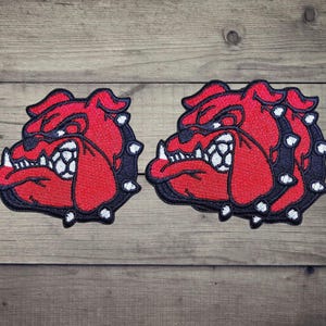 Può includere: Due toppe ricamate che raffigurano la testa di un bulldog di profilo. Il design utilizza filo rosso per il muso, filo nero per il collare e i contorni, e filo bianco per i denti e le borchie del collare, creando un aspetto grafico audace.