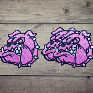 Pode incluir: Três emblemas bordados apresentando uma cabeça de bulldog estilizada em tons de rosa, preto e branco. Cada emblema retrata um bulldog rosnando com dentes proeminentes, um nariz escuro e um contorno preto espesso, com detalhes de costura.