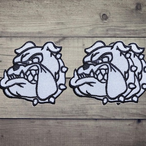 Pode incluir: Dois emblemas bordados com um design estilizado de cabeça de bulldog. Cada emblema é feito com fio branco e contornos pretos marcados, destacando as características faciais do cão, incluindo um focinho enrugado, orelhas pontiagudas e uma expressão determinada.