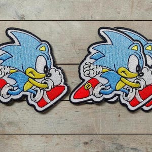 May include: Three embroidered patches featuring the character Sonic the Hedgehog in a running pose. The character has blue fur, a yellow chest, white gloves, and red shoes with gold buckles. Each patch is outlined in black thread with a white border.