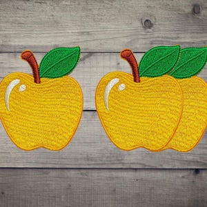 May include: Two yellow apple-shaped embroidered patches. Each apple features a brown stem, a single green leaf, and a white curved highlight on the left side of the fruit. The embroidery shows a dense, textured stitch pattern throughout the design.