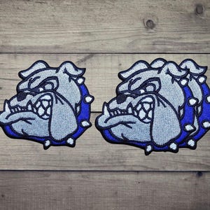 Può includere: Due toppe ricamate raffiguranti una testa di bulldog stilizzata in tonalità grigio chiaro e bianco, delineate da un bordo blu intenso. Il design presenta denti prominenti, un muso rugoso e un collare borchiato di forma circolare.