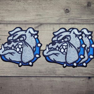 Peut inclure: Trois écussons brodés représentant la tête d'un bouledogue de profil. Le motif utilise du fil gris clair pour le visage, bleu foncé pour le contour et le collier, et blanc pour les dents et les pointes du collier, créant un visuel détaillé.