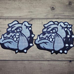 Puede incluir: Dos parches bordados idénticos que muestran una cabeza de bulldog estilizada de perfil. El diseño utiliza hilo azul claro para la cara y azul marino oscuro para los contornos y el collar, con detalles en blanco en los dientes y los ojos.