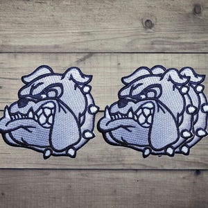 Puede incluir: Dos parches bordados que presentan una cabeza de bulldog estilizada de perfil. El diseño utiliza hilo azul claro y azul oscuro para delinear los rasgos faciales, incluyendo un hocico arrugado, orejas puntiagudas y dientes afilados.