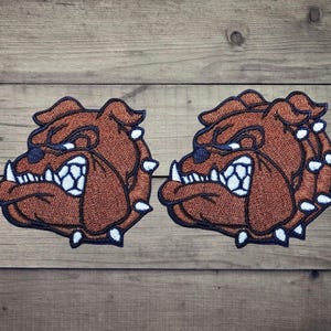 Puede incluir: Dos parches bordados idénticos que muestran la cabeza de un bulldog de perfil. El diseño utiliza hilo marrón para el pelaje, azul oscuro para los contornos y rasgos faciales, y hilo blanco para los dientes afilados y los colmillos visibles.