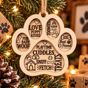 May include: A light-colored wooden paw-shaped ornament featuring engraved text and icons including Love Puppy Kisses, Zoomies, Fur Baby Woof, Best Friend, Playtime, Cuddles, Sniff, Fetch, and Treats, suspended by a rustic twine loop for hanging.