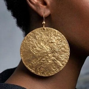Puede incluir: Un pendiente circular grande de color dorado con una superficie martillada muy texturizada. El pendiente cuelga de un sencillo gancho dorado, creando una apariencia metálica prominente y reflectante que capta la luz sobre su acabado irregular.