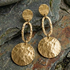 May include: A pair of gold-toned drop earrings featuring a hammered, textured finish. Each earring consists of a small circular stud connected to a vertical oval link, which suspends a larger circular disc at the bottom, creating a tiered design.