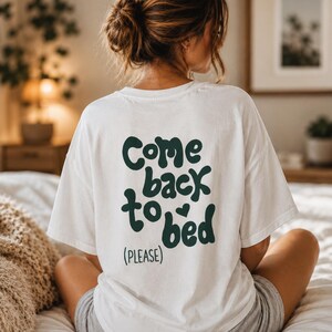 May include: A white short-sleeved t-shirt featuring dark green text on the back that reads "Come back to bed (PLEASE)" with a small heart icon above the word "bed". The shirt has a crew neckline and a relaxed fit, shown from the back view.