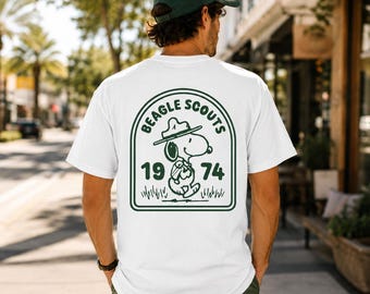 May include: A white t-shirt featuring a dark green graphic on the back. The design shows a cartoon beagle wearing a scout hat and backpack, framed by an arch with the text "BEAGLE SCOUTS" and the numbers "19" and "74" on either side.