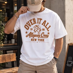 以下が含まれることがあります： OVER IT ALL 1996 Alleyway Crew NEW YORK GOOD PEOPLE BETTER DAYSという茶色のグラフィックプリント、ドラゴンのイラスト、星が描かれた白い半袖Tシャツ。ベージュのキャップとカーキ色のショートパンツを着用。