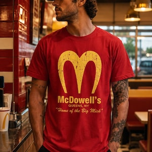 May include: A red short-sleeved t-shirt featuring a yellow distressed graphic of the letter M, with the text McDowell's QUEENS, NY and Home of the Big Mick printed in yellow below the logo on the front of the garment.