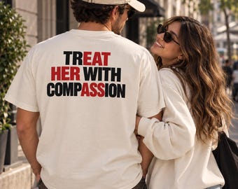 May include: An off-white short-sleeve t-shirt featuring the text TREAT HER WITH COMPASSION printed on the back. The word TREAT and the letters HER and COMPASSION are in black, while the letters HER and ASS are in red.